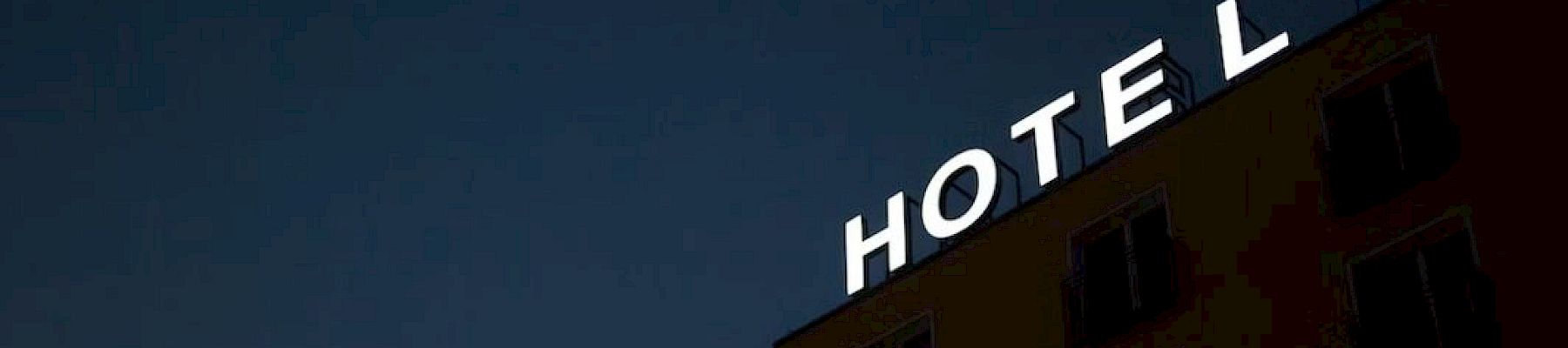 The image shows a building at night with an illuminated "HOTEL" sign missing the letter "L." There are several windows visible on the building.