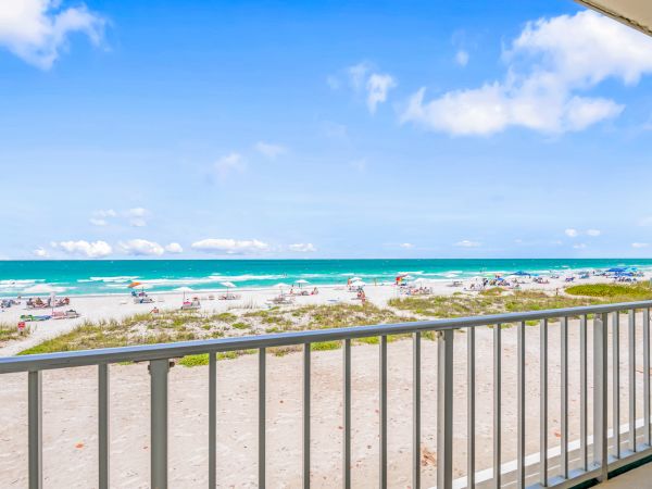 The image shows a sandy beach with people sunbathing and swimming, viewed from a balcony with a white railing under a clear blue sky.