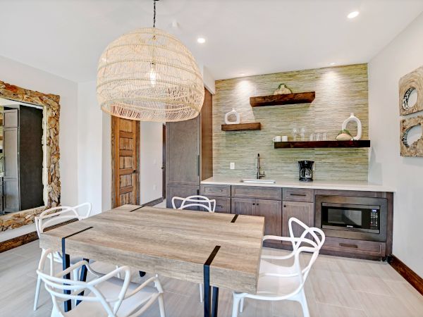 A modern dining area with a wooden table, white chairs, a large woven light fixture, a kitchenette, and decorative wall art and mirror.
