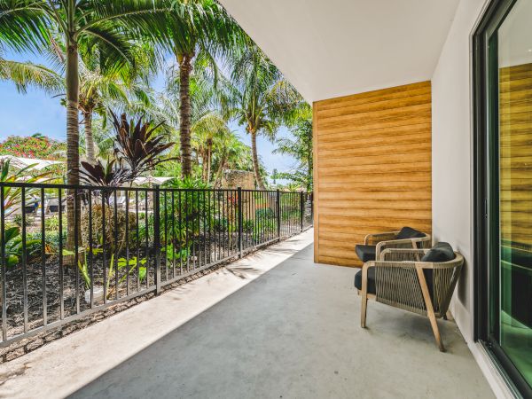 A sunny outdoor patio with two chairs, a railing, tropical plants, and a sliding glass door reflecting the greenery.