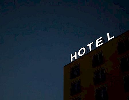 The image shows a building at night with an illuminated "HOTEL" sign missing the letter "L." There are several windows visible on the building.