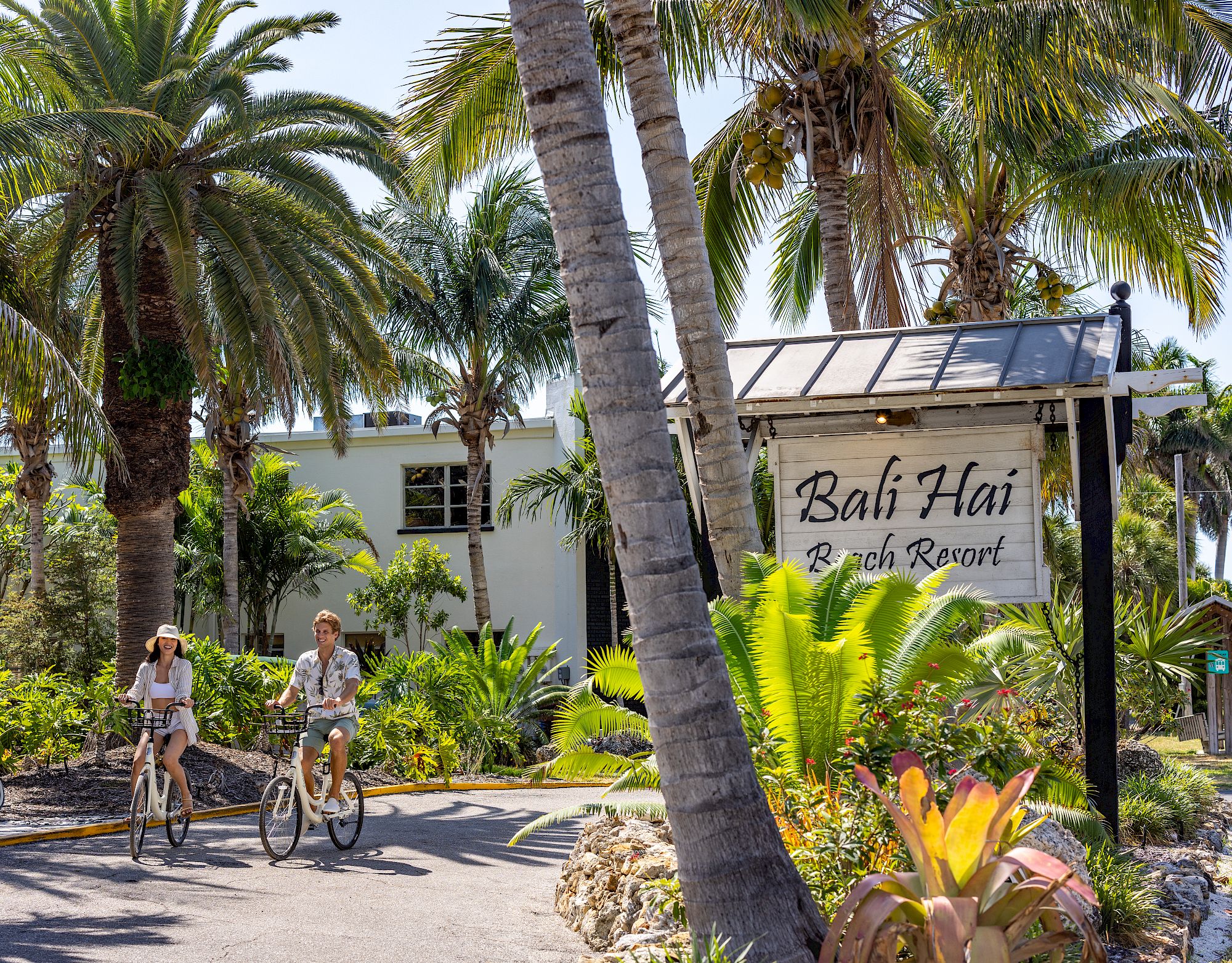 Two people ride bicycles near a tropical resort sign that reads "Bali Hai Beach Resort." Palm trees and lush greenery surround the area.