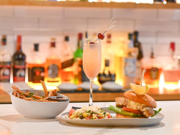 The image shows a bar counter with a drink, a bowl of fries and a burger with a side salad, set against a background of illuminated bottles.
