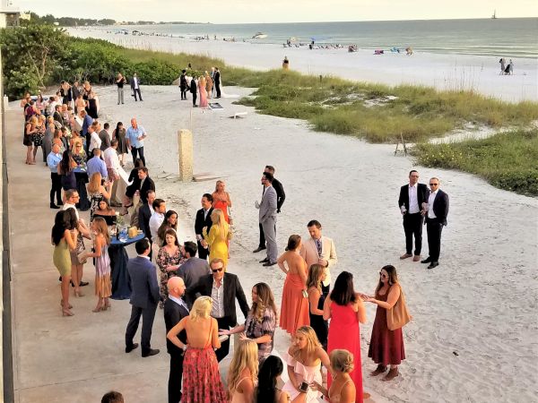 People in formal attire are gathered on a beachside promenade, mingling and enjoying what appears to be a social event or celebration.