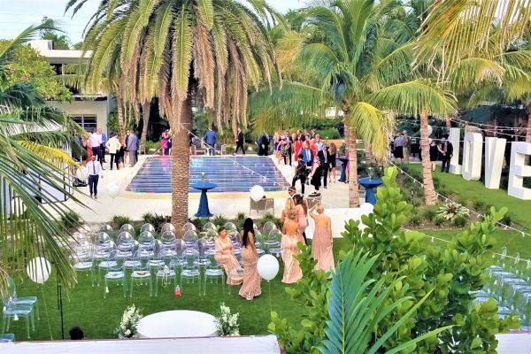 A lush outdoor setting features a pool, chairs, guests in formal attire, and large "LOVE" letters, indicating a wedding or event.