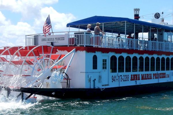 A red and white paddlewheel riverboat named "Anna Maria Princess" is cruising on the water with an American flag on its stern.