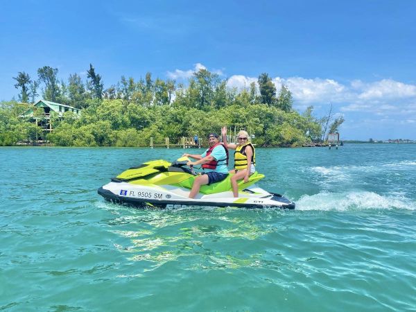 Two people riding a jet ski on a sunny day, with a lush island and trees in the background across a body of water.