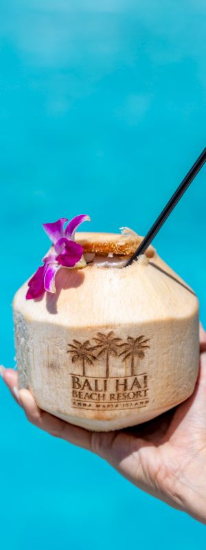 A hand holds a coconut drink adorned with a flower and straw against a blue water background. The coconut is labeled "Bali Hai Beach Resort and Spa."