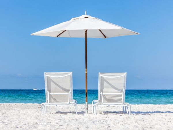 Two white lounge chairs and an umbrella on a sandy beach facing a calm blue sea under a clear sky.